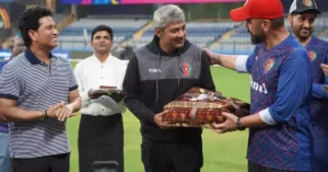 The image shows a moment during a cricket event, where former Indian cricketer Sachin Tendulkar, along with others, presents a gift to Afghanistan's team mentor. The gift exchange is happening on the field, with Tendulkar smiling and shaking hands with the mentor, who is wearing a black hoodie with the Afghanistan team logo. There are several people in the background, including a server holding an offering. The scene captures a warm and respectful exchange during a sporting event.